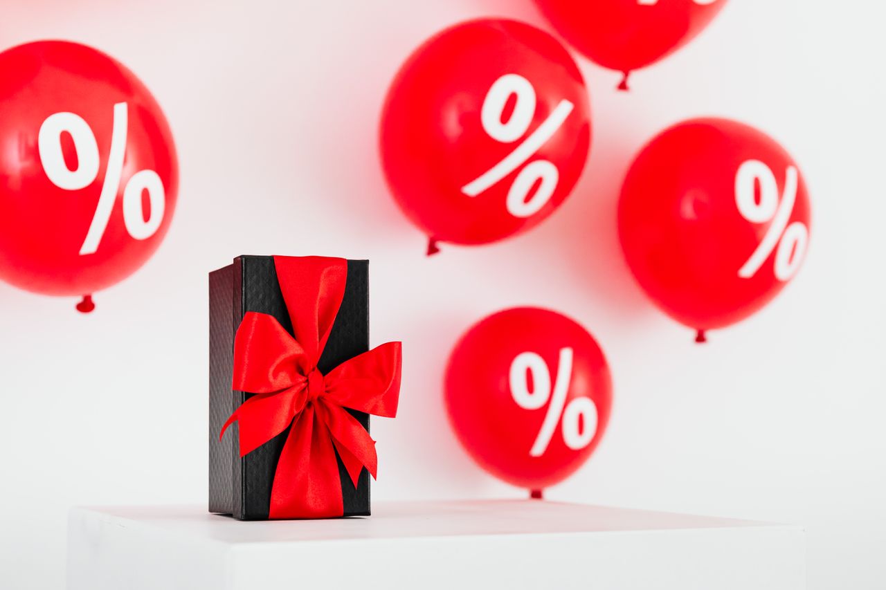 hero-01 Stylish black gift box with red ribbon surrounded by red percentage balloons on a white background.